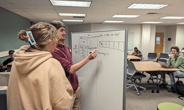 Students collaborate around a whiteboard in a classroom while discussing and solving a problem as others work nearby.