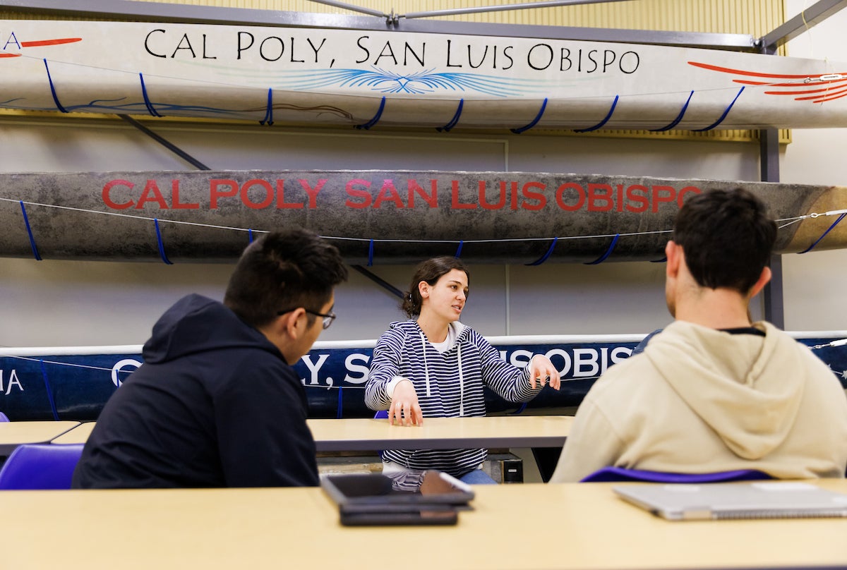 A female student in a striped hoodie speaks animatedly across a table to two fellow students in a Cal Poly San Luis Obispo classroom, with several branded concrete canoes mounted on the wall behind her.