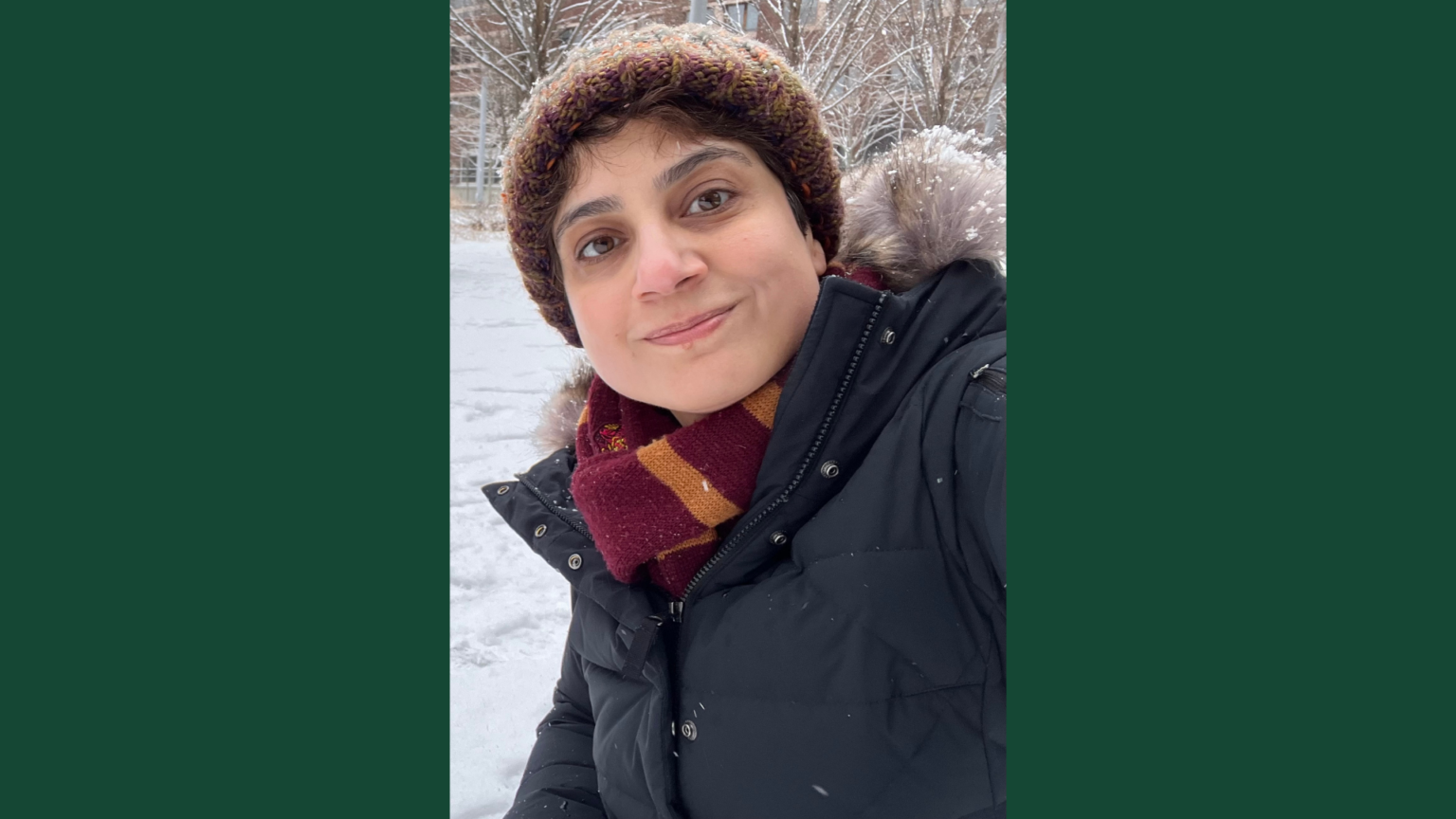 A woman smiles warmly at the camera in a winter selfie, wearing a knit beanie, a striped burgundy and gold scarf, and a black puffer jacket, with a snowy outdoor scene and bare trees visible behind her.