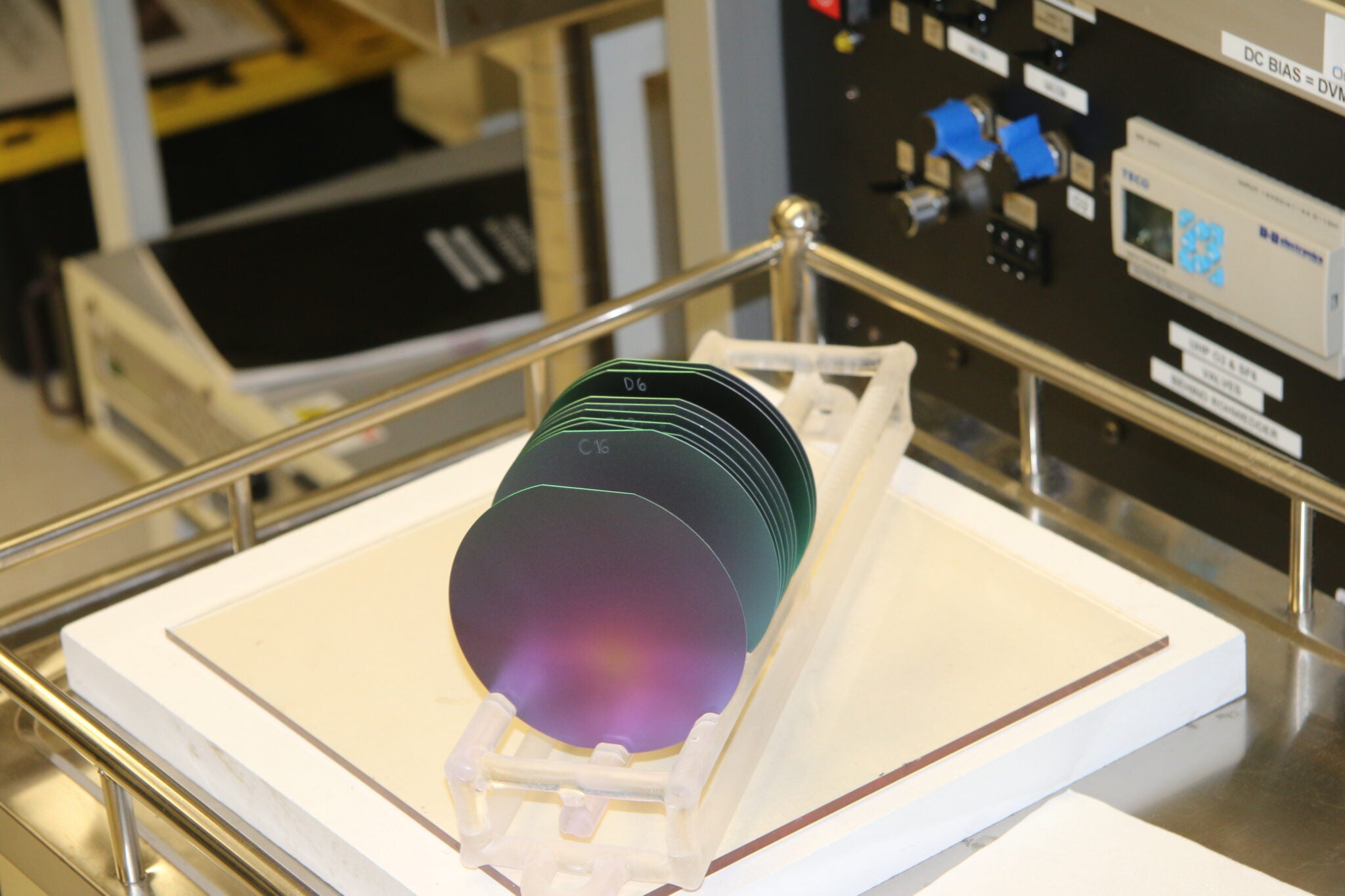 Several iridescent silicon wafers with colorful thin-film coatings stand upright in a transparent carrier rack on a lab bench, with fabrication equipment visible in the background.