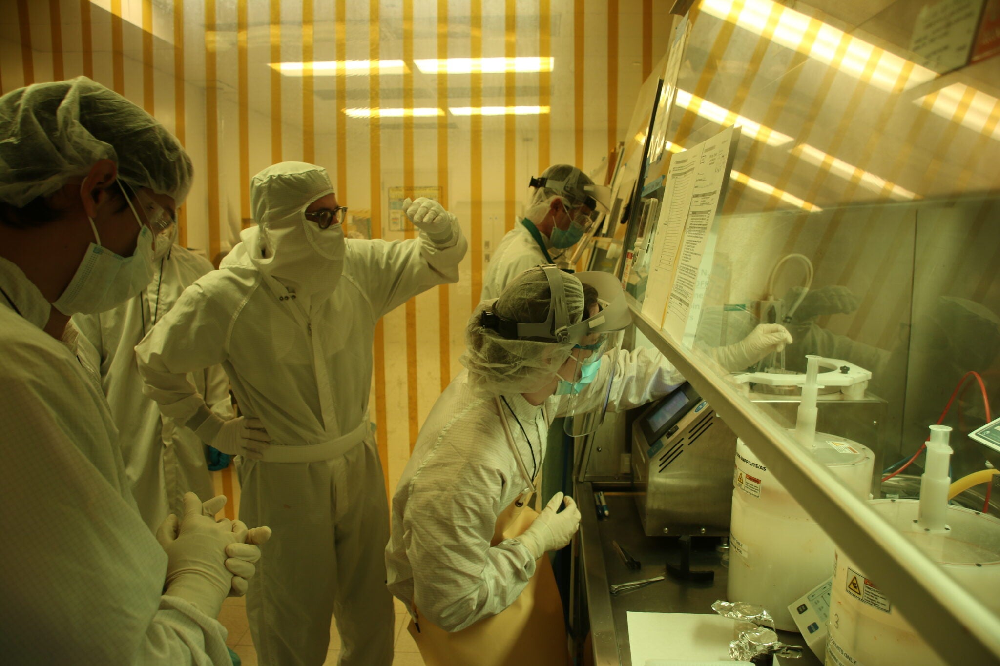 Multiple researchers in cleanroom suits, face shields, and gloves collaborate around a fume hood workstation in a yellow-lit microfabrication laboratory.