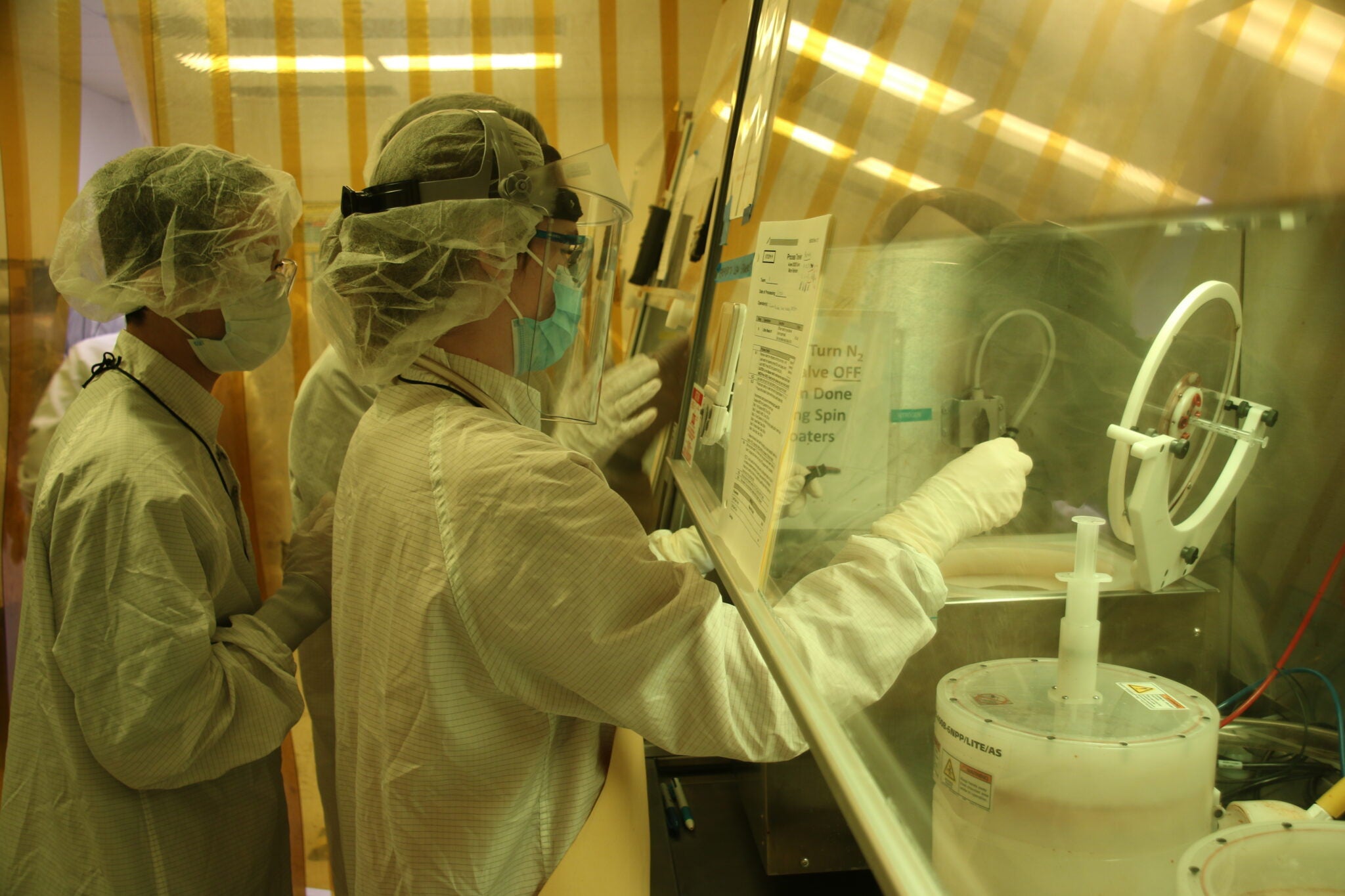 Two researchers in full cleanroom protective gear — including face shields, masks, hair nets, and gloves — work at a chemical fume hood in a yellow-lit microfabrication laboratory.
