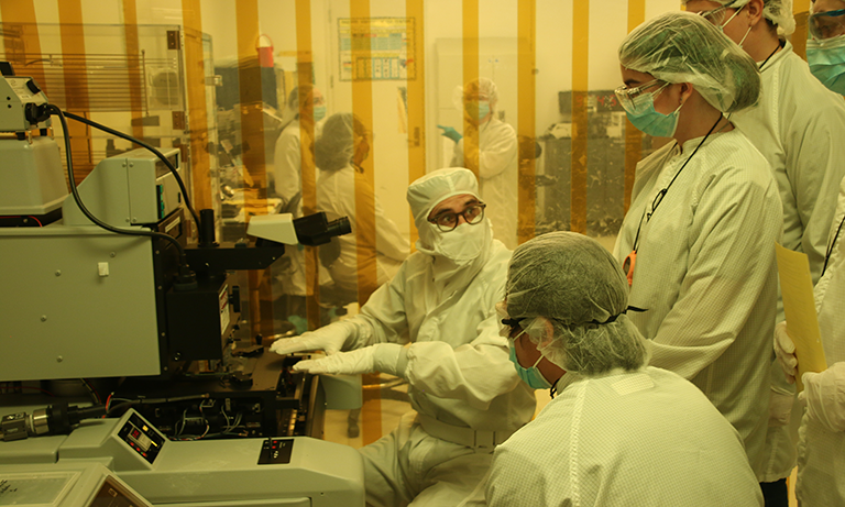 A group of cleanroom-suited researchers crowd around a large microscope or lithography machine in a yellow-lit semiconductor fabrication lab.