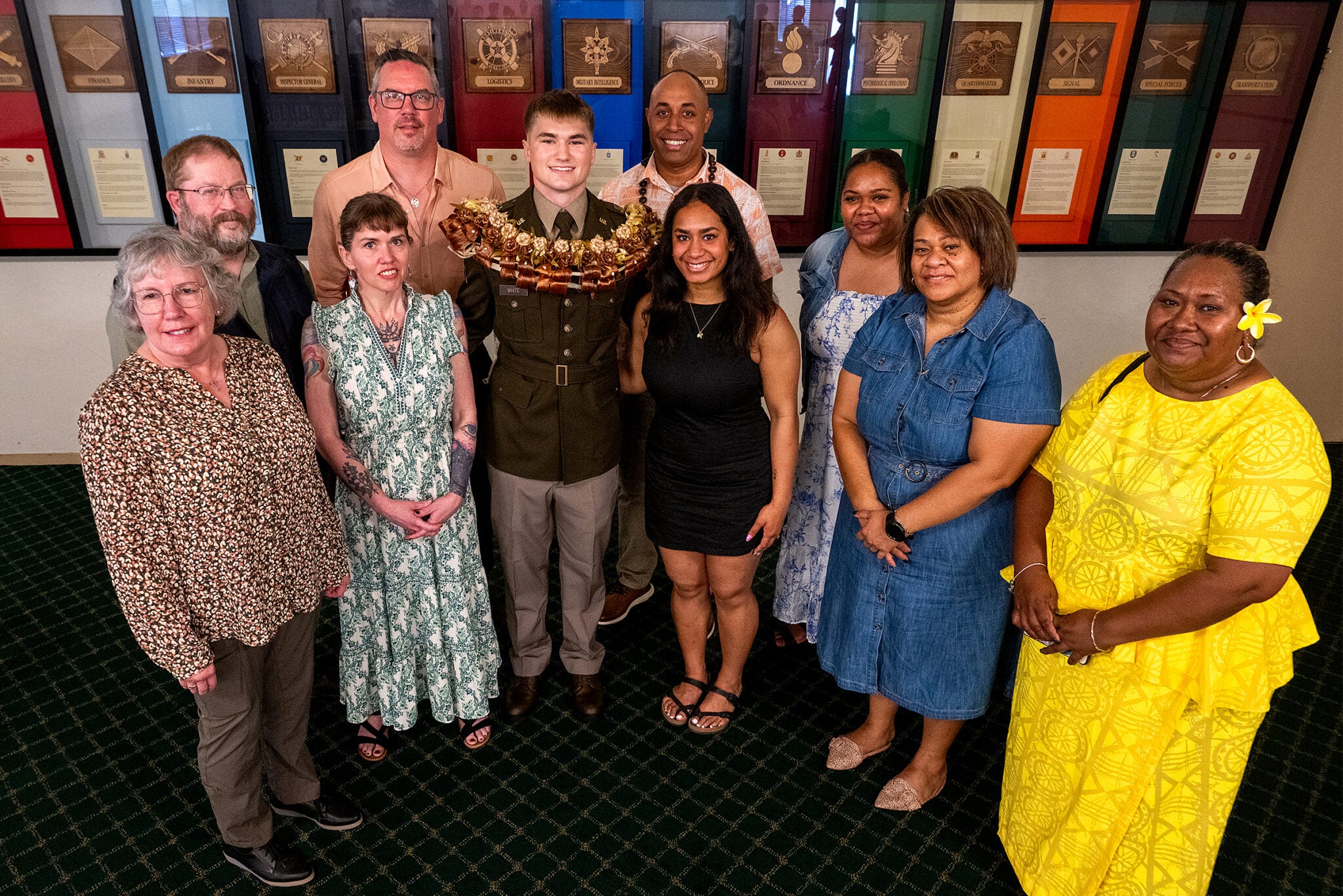 A newly commissioned Army officer named White, adorned with a large floral lei, poses with a group of eight smiling family members in front of military branch insignia plaques at the ROTC commissioning ceremony.