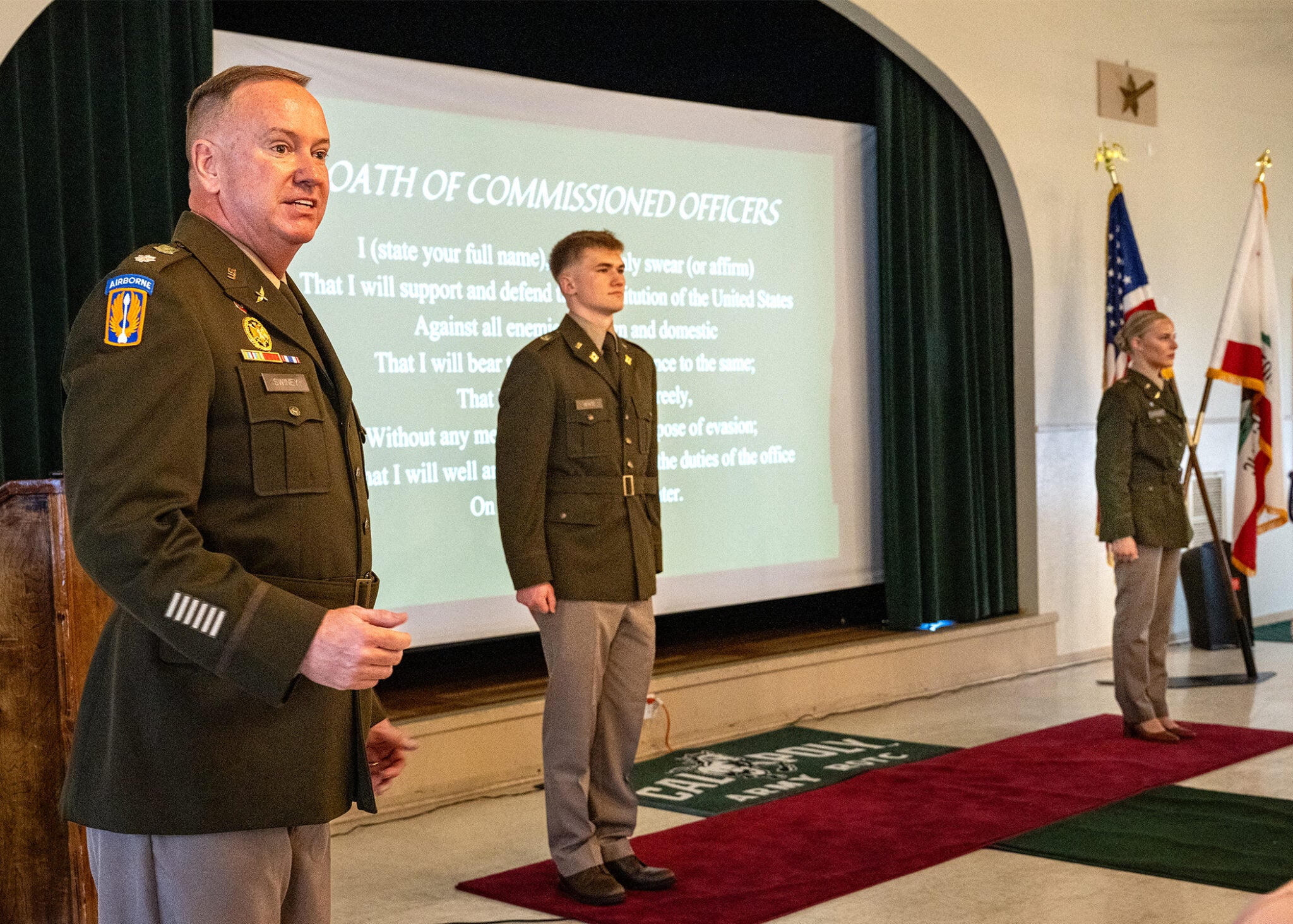 A senior Army officer in dress uniform leads the Oath of Commissioned Officers ceremony at Cal Poly's ROTC commissioning event, with two newly commissioned officers standing at attention before a projected slide of the oath.