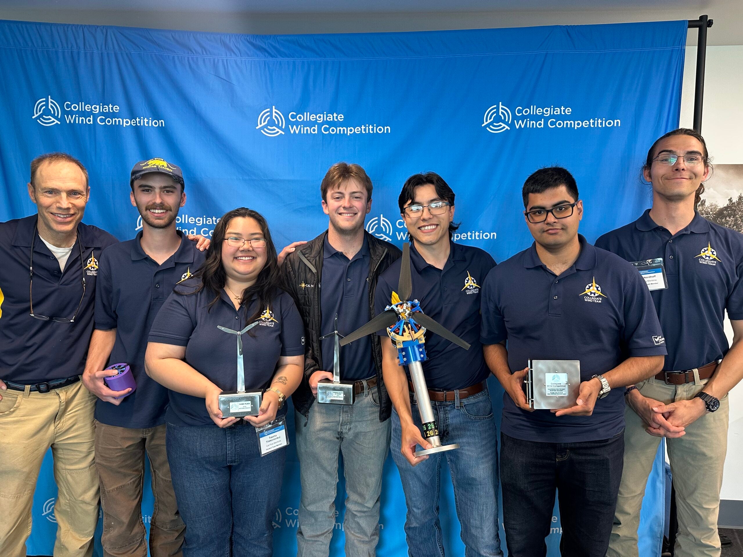 Seven members of the Cal Poly Collegiate Wind Team pose in matching navy polos in front of a Collegiate Wind Competition backdrop, holding trophies and their small-scale wind turbine model.