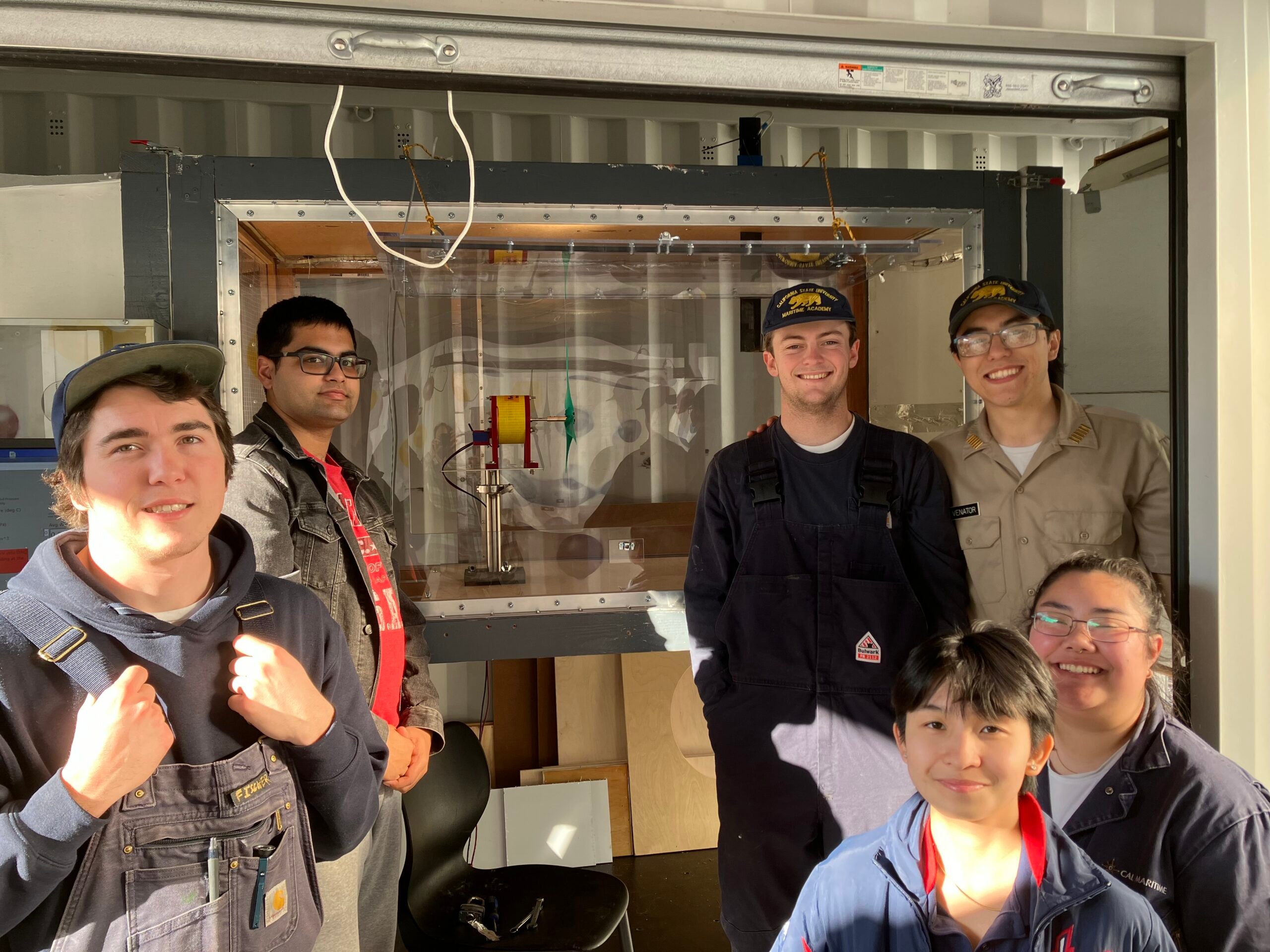 Six Cal Maritime students smile in front of a wind tunnel containing their small-scale wind turbine prototype, with some wearing their maritime academy uniforms.