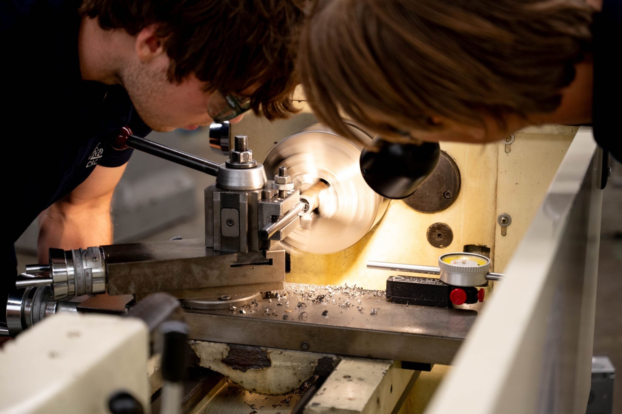 Two students lean in closely to observe a spinning metal component being precision-machined on a metal lathe, with metal shavings visible on the machine bed.
