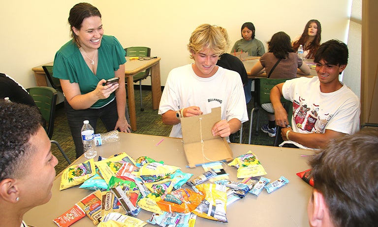 Professor times students using a "robot arm" made from cardboard to pick up snack packets.