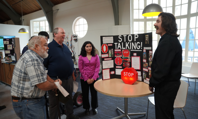 A student standing in front of his project, which includes a giant red stop sign. He is explaining his project to bystanders.