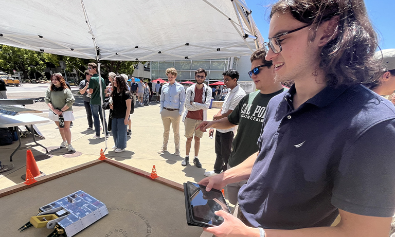 A student holding a tablet that is controlling a device in front of him, while other students look on in the background