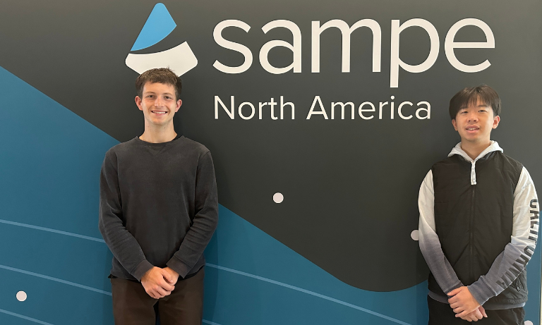 Two students standing in front of a wall that has SAMPE North America printed on it