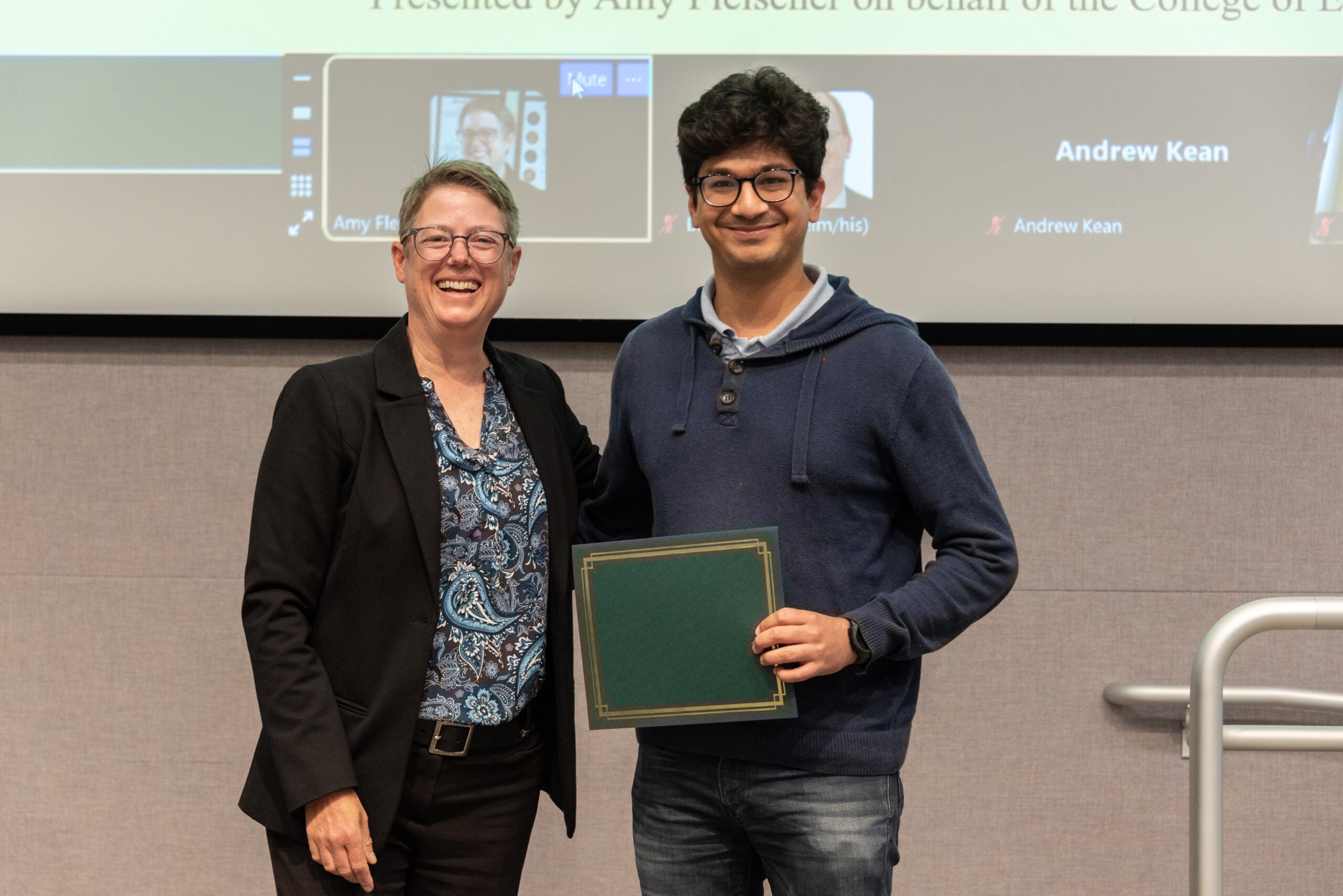 Two people standing smiling at the camera, with the person on the right holding up a paper certificate