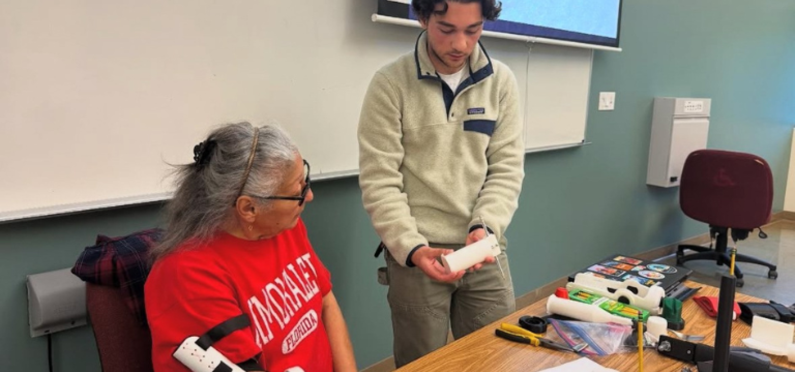 A student holding part of a 3D printed prosthesis, while a woman sitting to his right tries on another part of the prosthesis