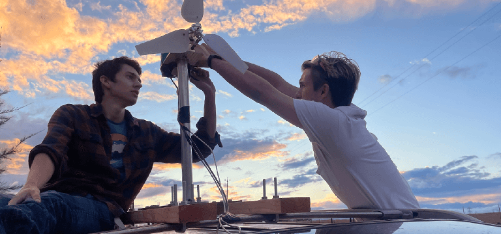 Two students sitting on top of a car, working on a wind turbine