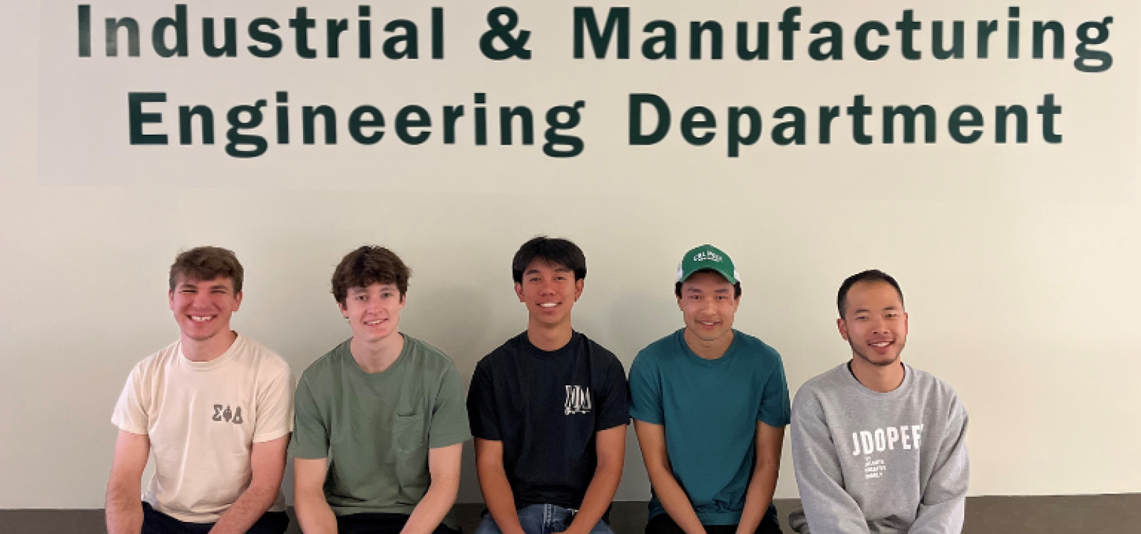 Five students sitting down and smiling while facing the camera. They are sitting in front of a wall that has "Industrial & Manufacturing Engineering Department" on it.