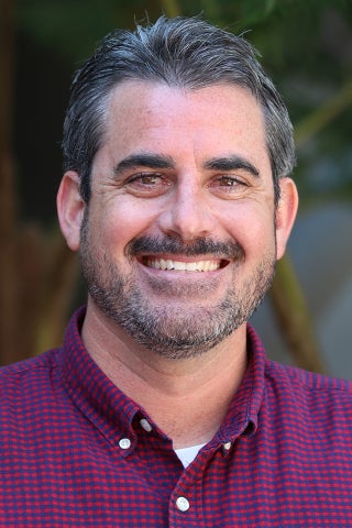 Headshot of a smiling man with dark salt-and-pepper hair and a beard, wearing a red and navy checkered shirt, photographed outdoors with a blurred green background.