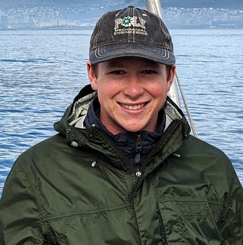A smiling young man wearing a Cal Poly Mechanical Engineering baseball cap and a green L.L.Bean rain jacket, photographed outdoors with a blue body of water and a cityscape in the background.