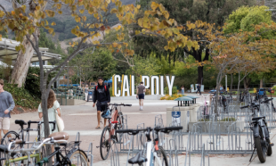 Campus walkway at Cal Poly with students walking and bike racks in the foreground, framed by trees and a large Cal Poly sign in the background.