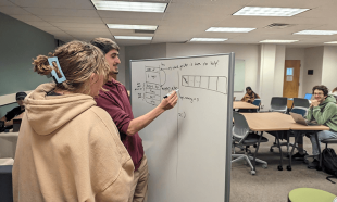 Students collaborate around a whiteboard in a classroom while discussing and solving a problem as others work nearby.