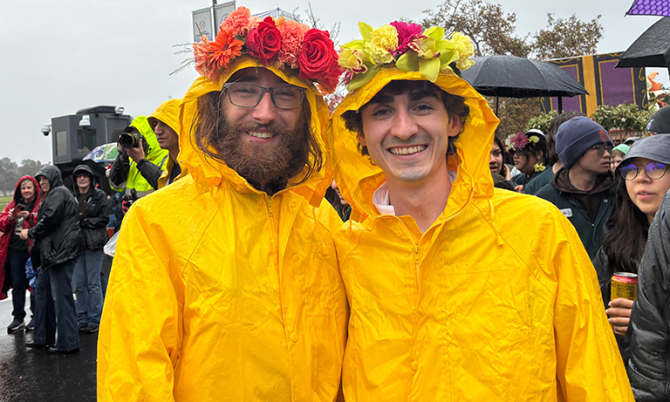 Two smiling students standing outside in a gentle rain, both wearing vibrant yellow raincoats and flower crowns.