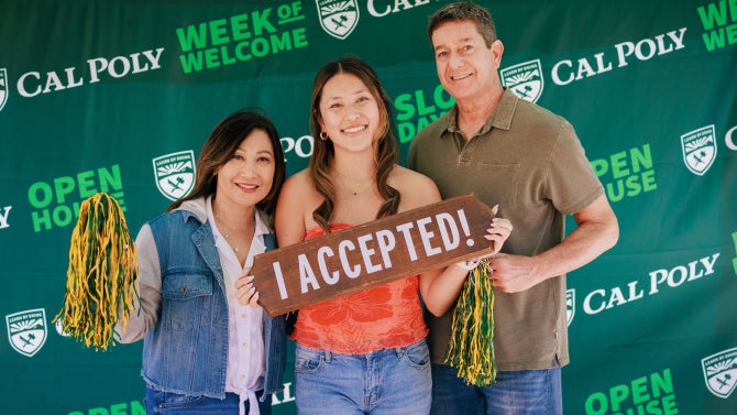 group of 3 people standing in front of Cal Poly banner holding props