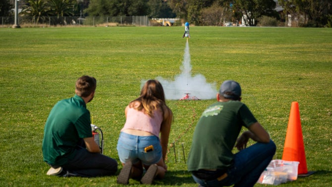 Aerospace Engineering rocket competition, three students launching rocket on grass field