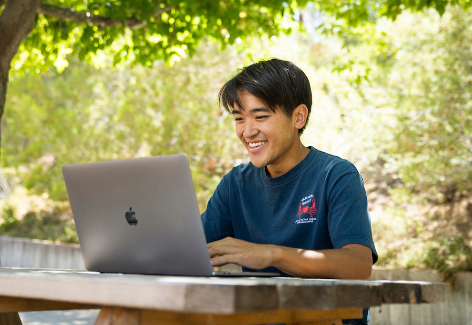 Smiling student working on a laptop while sittin on a bench