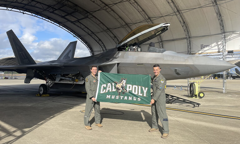 Two fighter pilots stand by an F-22 holding a Cal Poly banner