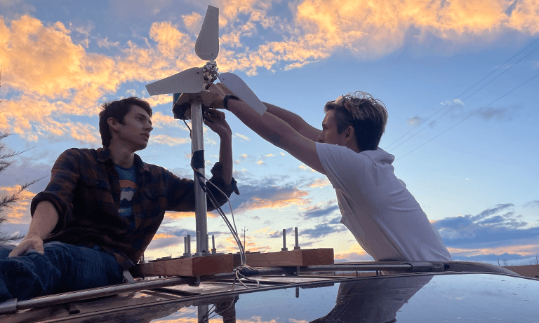 Two students sitting on top of a car, working on a wind turbine