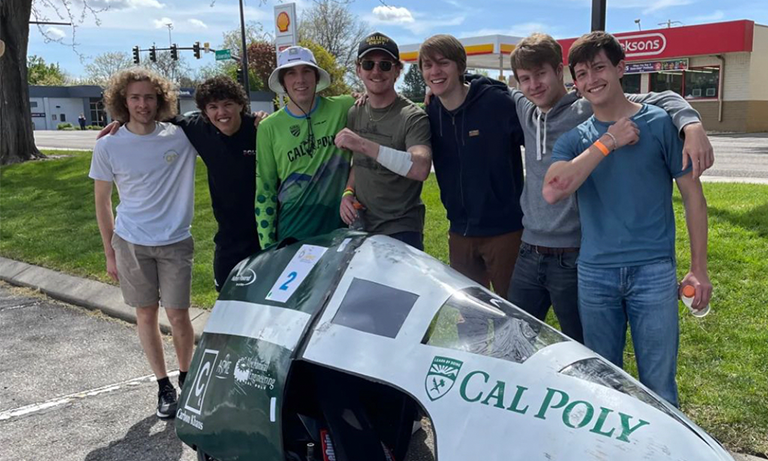 Seven students posing with their arms around each others' shoulders and a racing vehicle in front of them