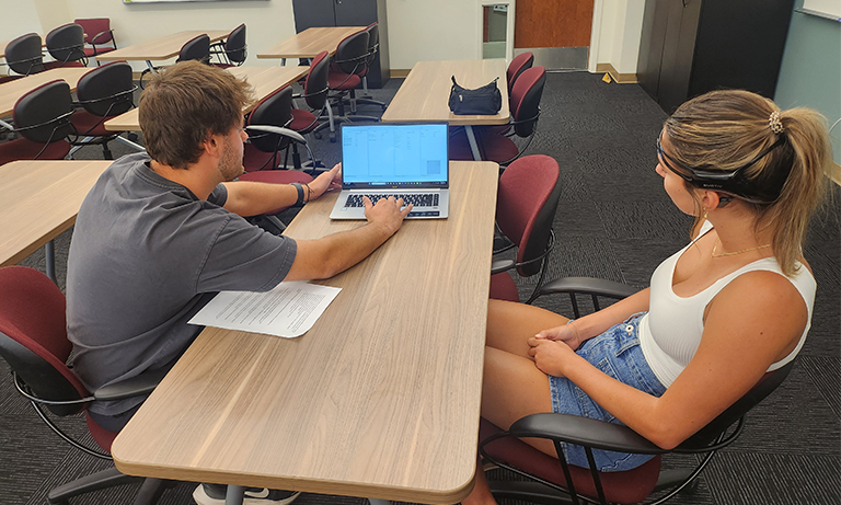 A man looking at a computer and a woman sitting across from him with a headset on.