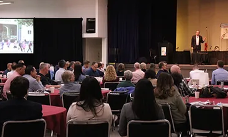 A group of people sitting at tables during a conference and watching the speaker on stage.