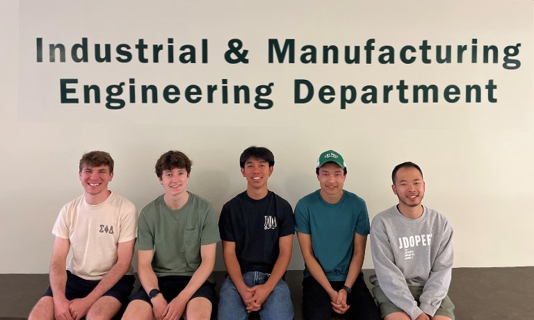 Five students sitting down and smiling while facing the camera. They are sitting in front of a wall that has "Industrial & Manufacturing Engineering Department" on it.