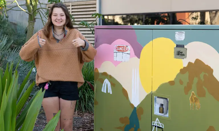 A person holding two thumbs up in front of a utility box that they painted.