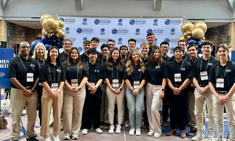 Students and faculty members posing for a group photo in front of a step and repeat banner at a conference.