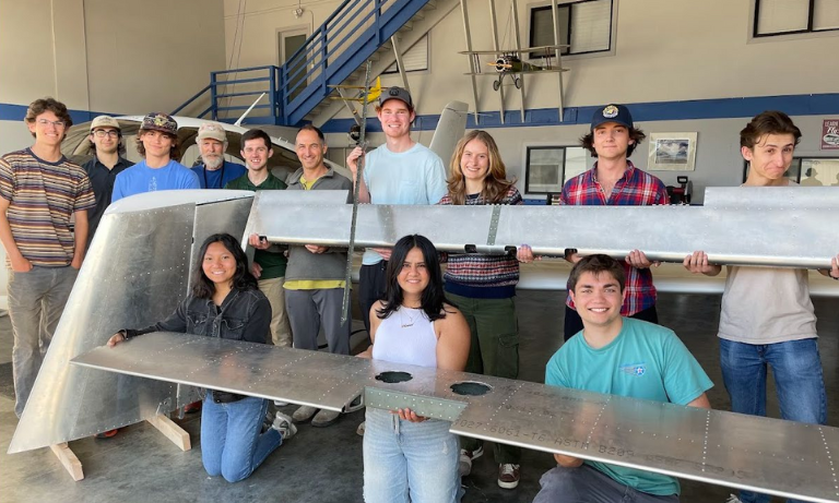 A group of students and faculty members holding up parts of a plane and smiling at the camera.