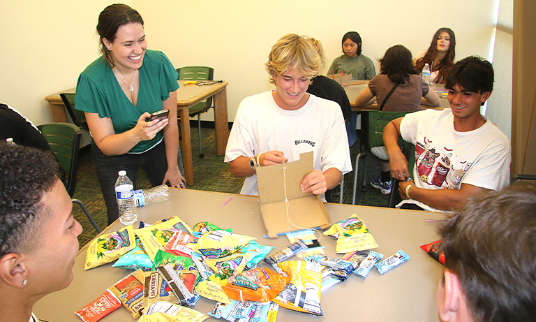 Professor times students using a "robot arm" made from cardboard to pick up snack packets.