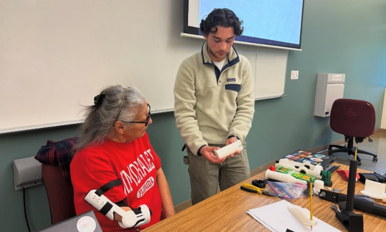 A student holding part of a 3D printed prosthesis, while a woman sitting to his right tries on another part of the prosthesis