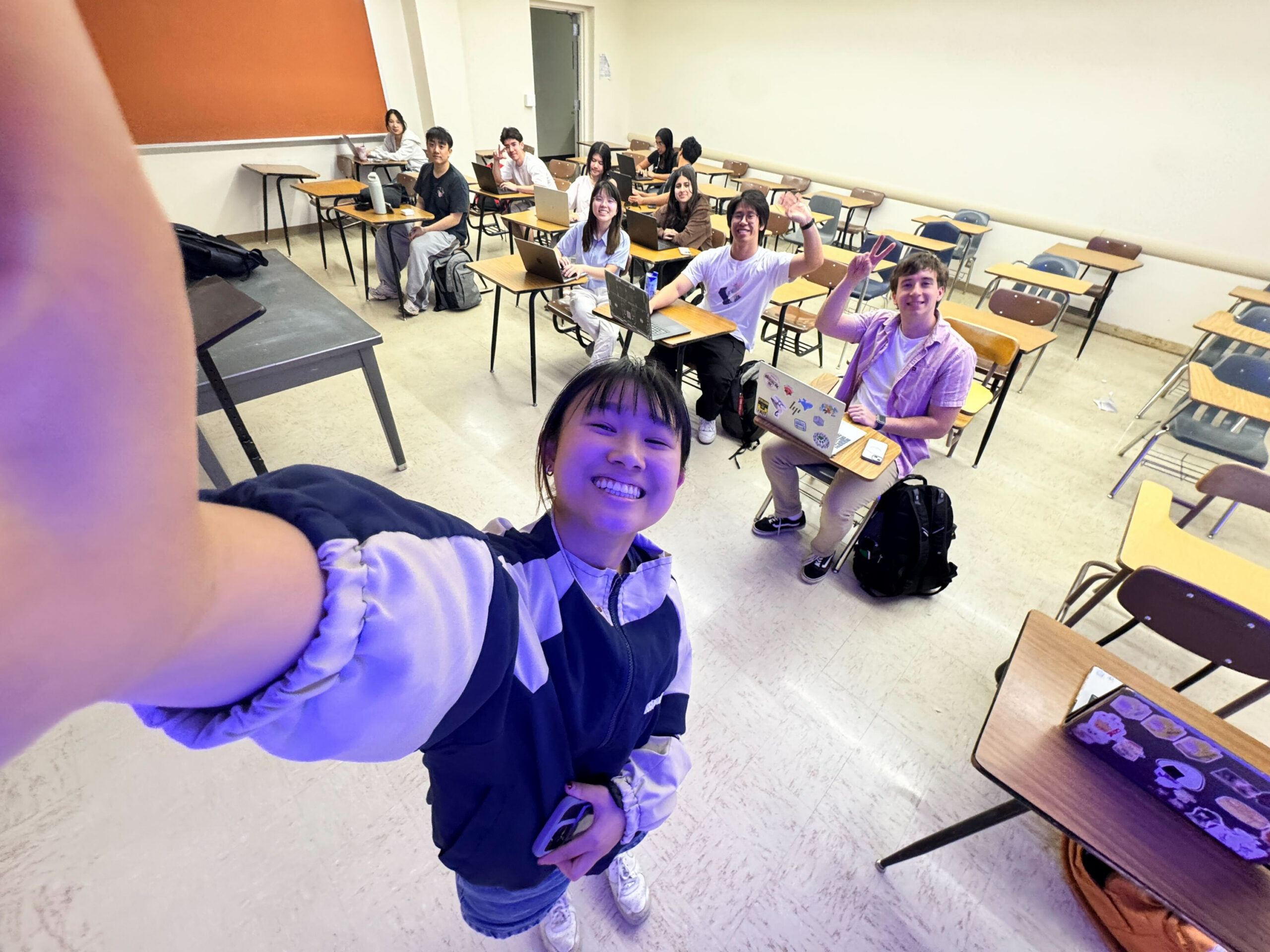 Student takes selfie with her team in the background of a classroom, working on a project